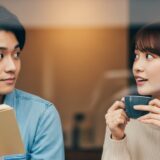 フェの窓際で、二人の視線が偶然交差する瞬間。男性は本を読んでいて、女性はコーヒーカップを持っている。お互いに少し驚いたような、でも嬉しそうな表情
