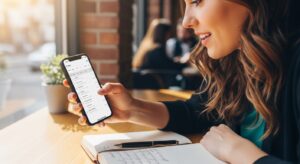 スマートフォンでデートプランを確認している女性の手元。カフェのテーブルの上にノートとペン、スマホが置かれている。事前準備をしているイメージ。