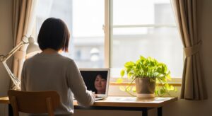 窓際の明るいデスクでノートパソコンに向かう日本人女性の後ろ姿。自然光が差し込み、観葉植物も置かれている。