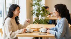 カフェで向かい合って話をしている二人の日本人女性。笑顔で会話している様子。友達との時間を表現。自然光が入る明るい雰囲気。