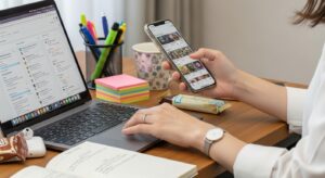 スマートフォンとノートパソコンを同時に操作している女性の手元。効率よく作業している様子。デスクには文房具やメモも置かれている。